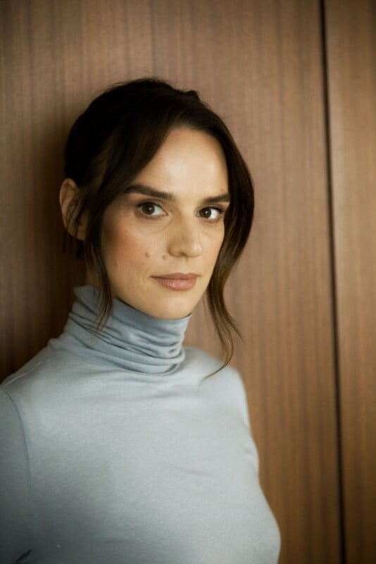 A woman with dark hair in a low updo, wearing a light gray turtleneck, stands in front of a vertical wooden panel background, looking directly at the camera with a neutral expression.