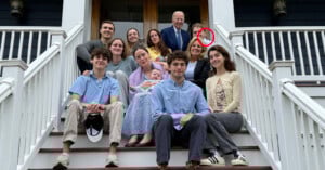 A multigenerational family of twelve, including a baby, poses and smiles together on the front steps of a house. A man in a suit stands at the top; a woman behind him is circled in red.