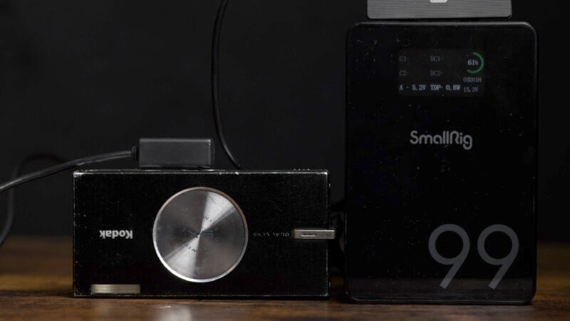 A close-up of a Kodak film camera next to a SmallRig device. The camera has a prominent round dial, and the SmallRig device displays a digital readout. Both are set on a wooden surface against a dark background.