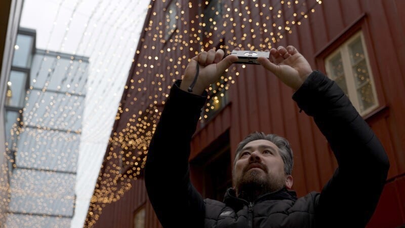 A person with a beard holds a camera above their head, capturing a scene of twinkling lights overhead. They stand between tall buildings with string lights creating a festive atmosphere. The person wears a dark jacket, looking upwards.