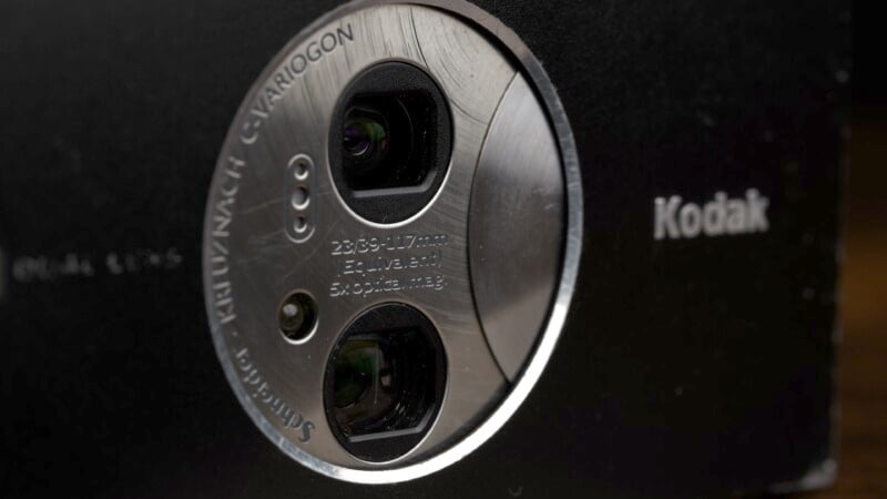 Close-up of a Kodak camera lens with visible details such as "24-86mm" and "f/2.8-4.9" specifications on a metal ring. The lens features dual openings and the brand name "Kodak" is visible on the camera body.