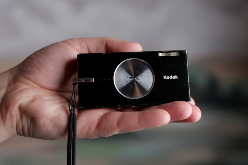 A hand holding a compact Kodak digital camera. The camera is black with a silver lens and has a wrist strap attached.