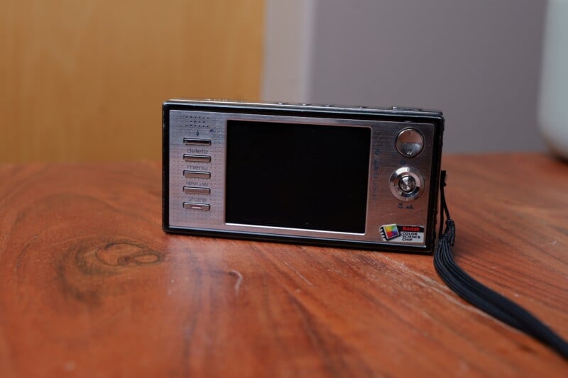 A sleek, black and silver digital camera with a large screen on a wooden surface. It features control buttons and a small joystick on the side, along with a Kodak logo at the bottom right corner. A wrist strap is attached to the camera.