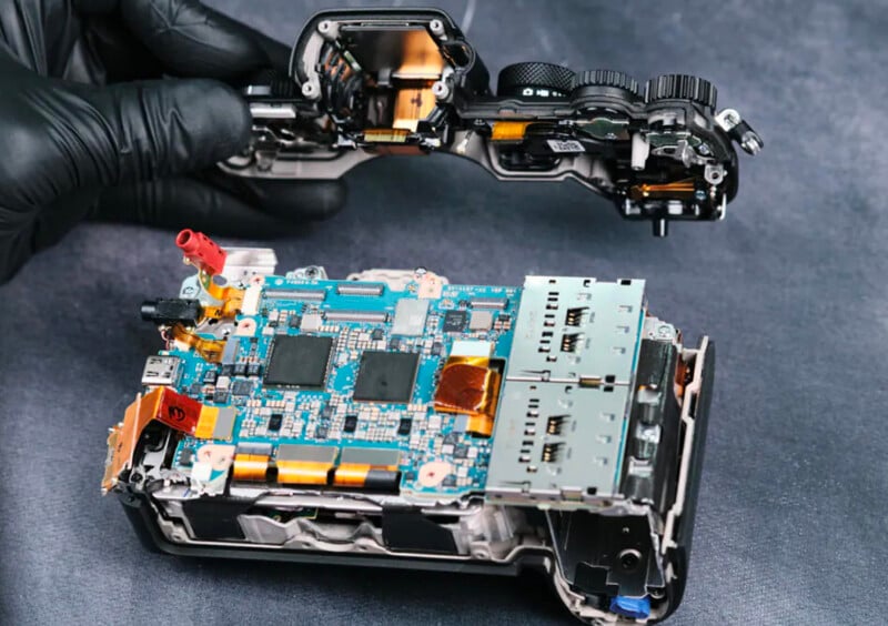 A gloved hand lifts the top cover off a digital camera, revealing its internal circuit boards, electronic components, and wiring on a dark surface.