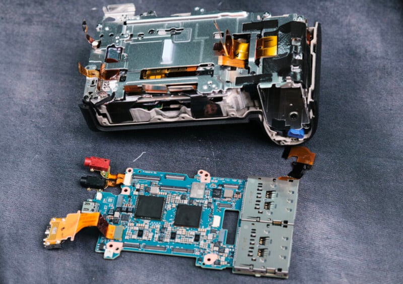 A disassembled electronic device on a dark surface, showing its internal circuit board and various components, including connectors, chips, and ribbon cables.