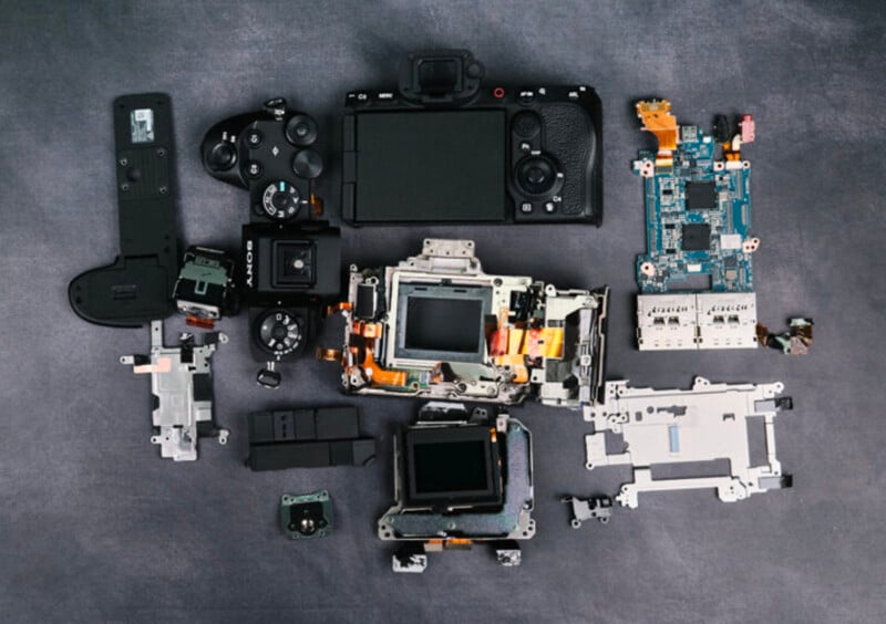 A digital camera is disassembled and laid out on a grey surface, showing various internal components such as circuit boards, lenses, screens, and metal frames.