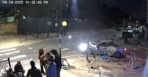 A nighttime street scene shows a group of people gathered on a sidewalk near film equipment. A silver car is parked by a zebra crossing, and bright lights illuminate the area. The timestamp reads “04-14-2025 10:32:43 PM.”.