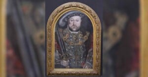 A framed painting of a man in ornate Tudor-era clothing, wearing a hat and holding a sword. The detailed gold frame is arched, and the background is dark, highlighting the subject’s elaborate jewelry and attire.