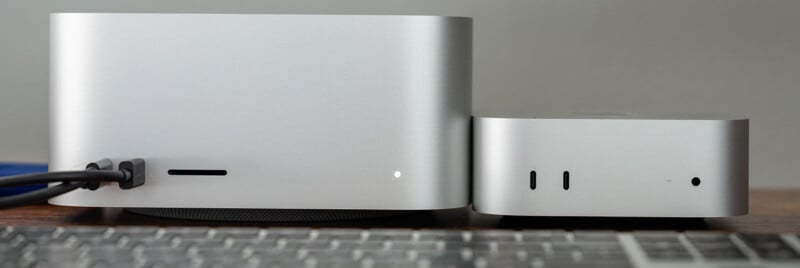 Two silver desktop computers are placed side by side on a wooden surface. The larger device features multiple ports and a power indicator, while the smaller one also has ports and indicators. A keyboard is partially visible in the foreground.