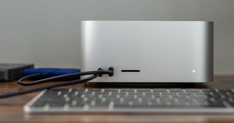 A silver computer on a wooden desk with cables plugged into its back. A keyboard is partially visible in the foreground, and a blue object is next to the computer. The computer has a small white light on its front.