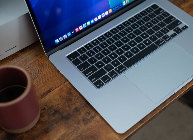 A laptop with a dark keyboard and open screen, displaying a desktop with app icons, is on a wooden table. Next to it, there's a maroon ceramic mug with a black liquid inside. A silver box is partially visible in the background.