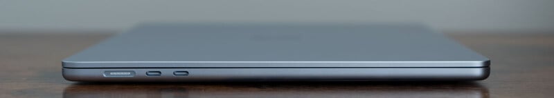 Side view of a closed, thin laptop on a wooden surface, showing two USB-C ports and a headphone jack. The laptop is sleek and metallic, with a minimalist design.
