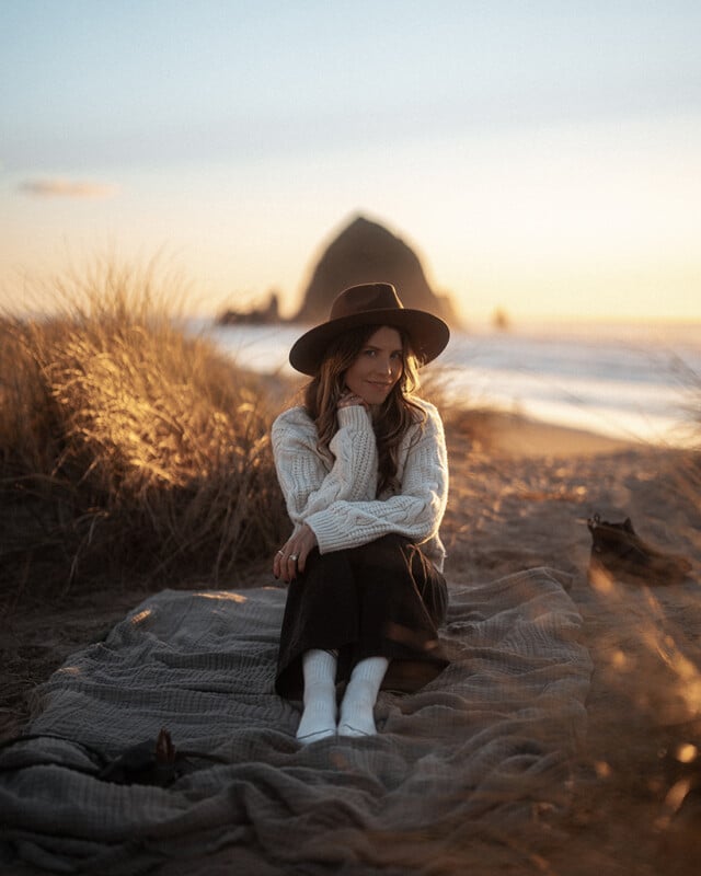 A woman wearing a hat and cozy sweater sits on a blanket in grassy sand dunes at the beach during sunset, with a large rock formation and ocean in the background.