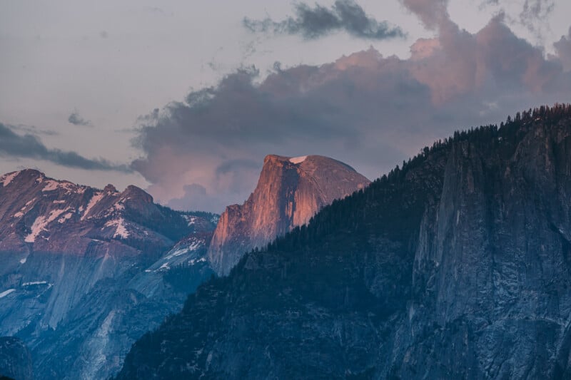 Half Dome mountain in Yosemite National Park is illuminated by the soft pink light of sunset, with dramatic clouds in the sky and snow patches visible on surrounding peaks.