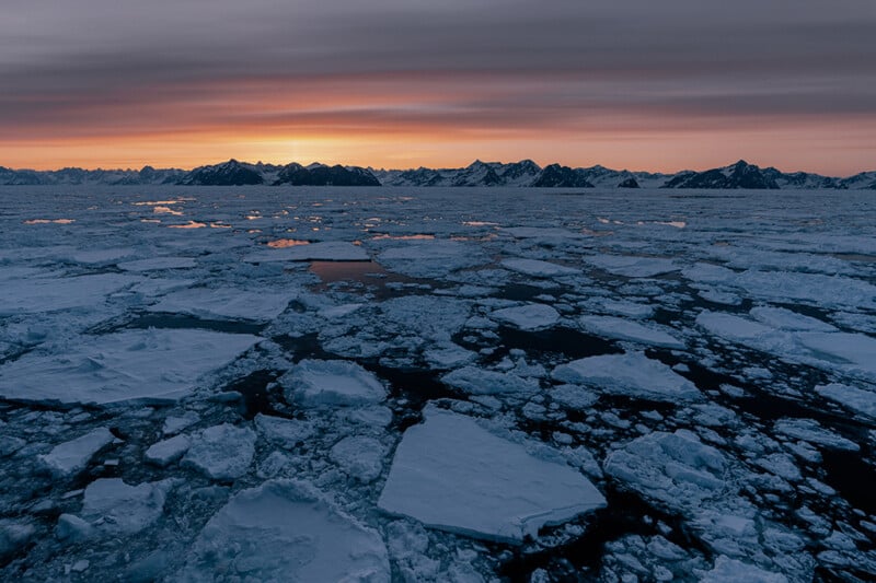 Large chunks of sea ice float on dark water under a dramatic sky at sunset, with distant mountains silhouetted on the horizon.