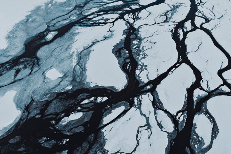 Aerial view of cracked ice on a frozen body of water, with dark cracks and channels forming abstract, branching patterns across the icy surface. The scene appears cold and stark.