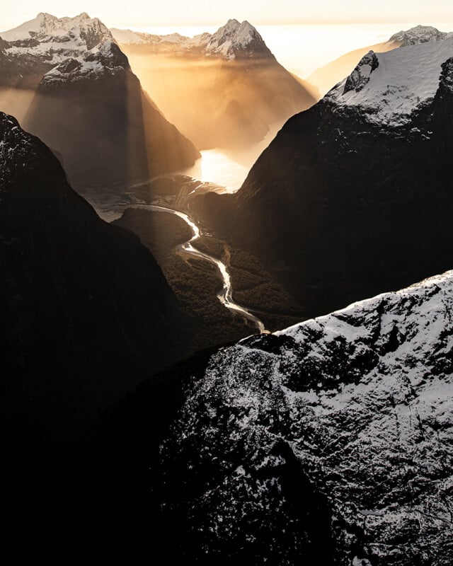 A river winds through a deep valley surrounded by snow-capped mountains, with golden sunlight streaming through clouds and illuminating the water and landscape below.