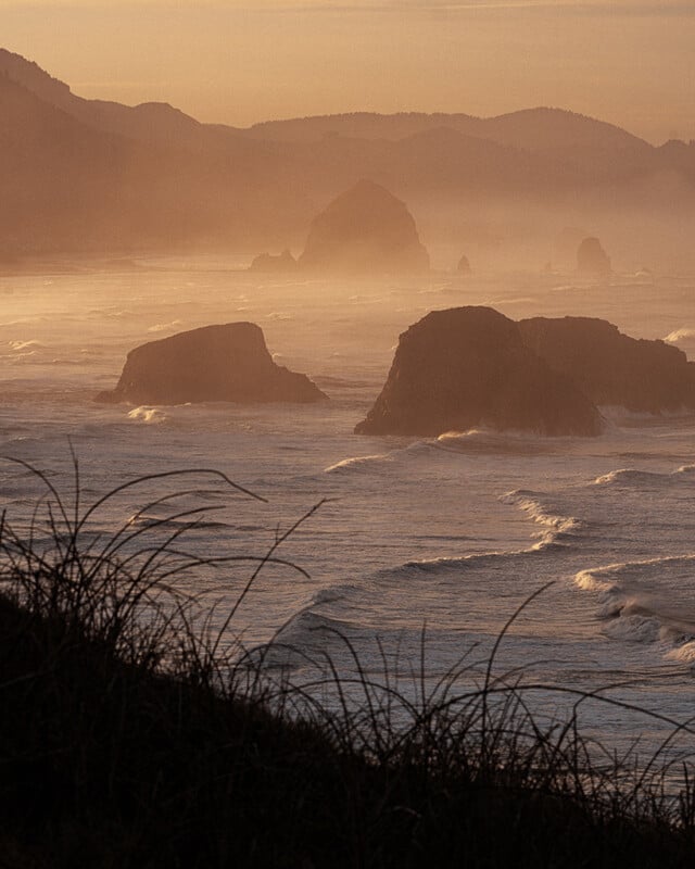 A coastal landscape at sunset, with sea stacks rising from the waves, mist drifting over the ocean, and silhouetted hills and grasses in the foreground.