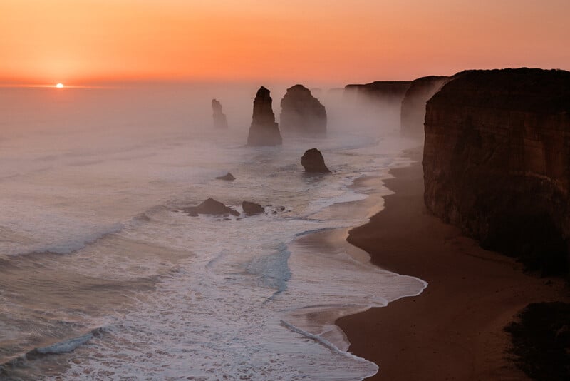 Sunset over the ocean with waves crashing against tall limestone rock formations and cliffs along a misty coastline, likely the Twelve Apostles in Australia.