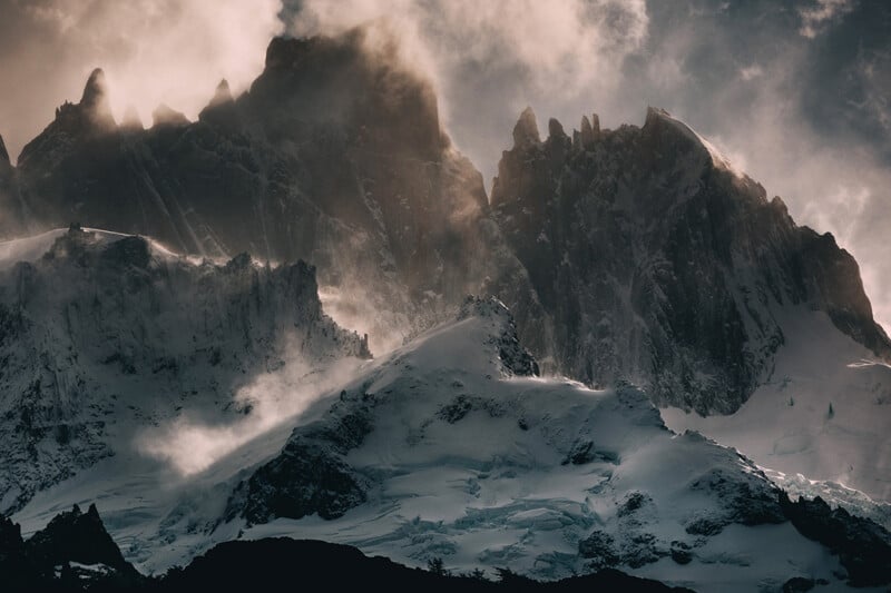 Dramatic mountain peaks covered in snow rise sharply into the clouds, with sunlight streaming through mist and fog, creating a moody and atmospheric scene.