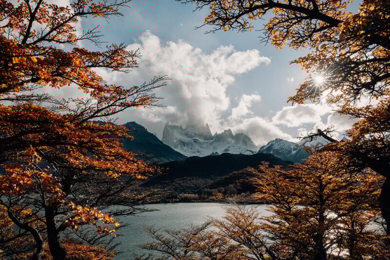 Snow-capped mountains framed by autumn trees with orange leaves, a lake in the foreground, and sunlight streaming through branches under a partly cloudy sky.