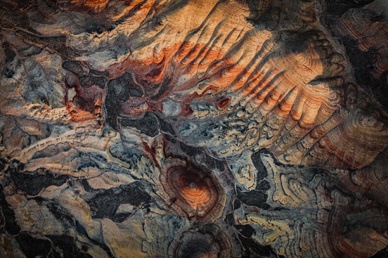 Abstract aerial view of a rugged landscape with swirling patterns in shades of orange, brown, gray, and black, resembling layers of rock or mineral formations illuminated by soft sunlight.