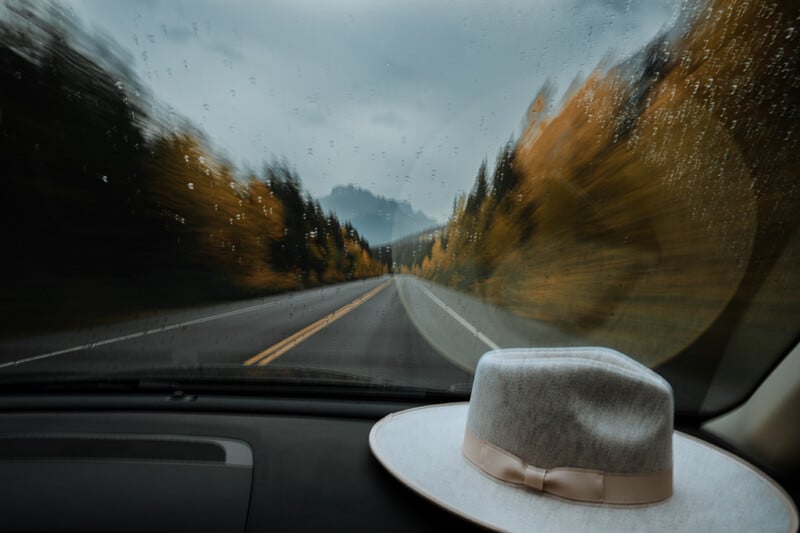 View from a car windshield on a rainy day, looking down an empty road lined with autumn trees. Raindrops blur the glass. A light gray hat with a ribbon sits on the dashboard.