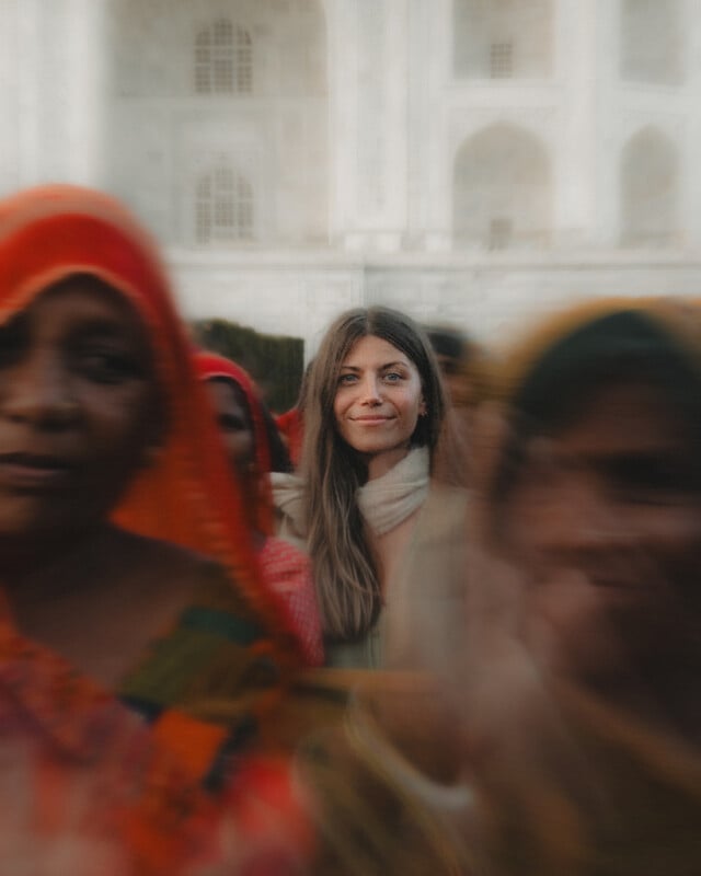 A woman with long brown hair and a light scarf stands smiling in focus, surrounded by blurred figures in colorful clothing, with a white building featuring arched windows in the background.