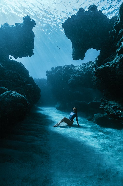 A woman in a swimsuit sits on the sandy ocean floor, surrounded by large, shadowy coral formations and illuminated by beams of sunlight filtering through the water above.