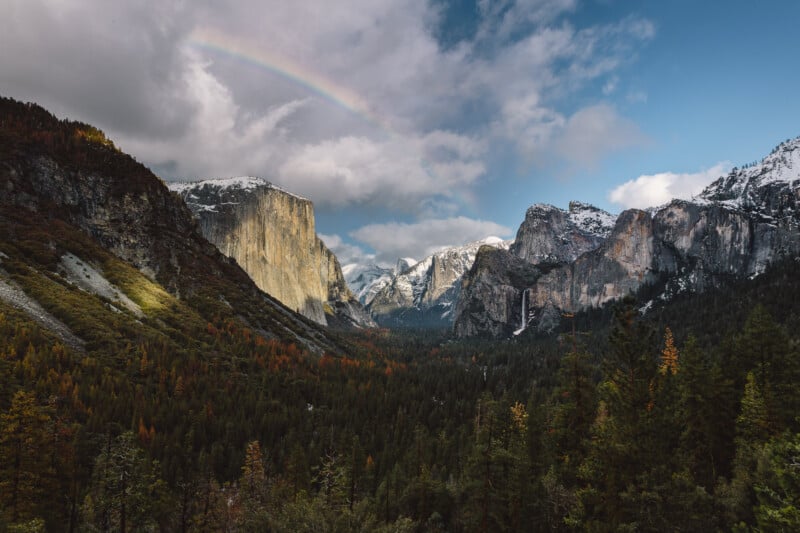 A dramatic view of Yosemite Valley with El Capitan on the left, Half Dome in the distance, Bridalveil Fall on the right, lush forest below, and a faint rainbow arching across a partly cloudy sky.