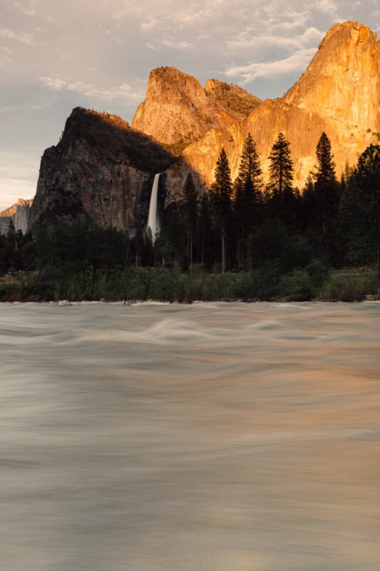 A waterfall cascades down a cliff surrounded by pine trees, with tall rocky mountains in the background glowing in the warm light of sunset. A river flows calmly in the foreground.