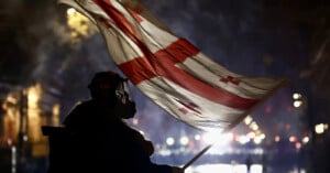 A silhouette of a person wearing a mask holds up a large flag with a red cross and four red smaller crosses, against a backdrop of city lights at night.