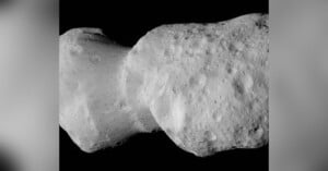 A close-up image of a gray, irregularly shaped asteroid in space, showing a rough, cratered surface with two distinct lobes connected at a narrow region against a black background.