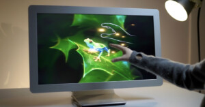 A person touches a computer monitor displaying a vivid image of a frog on green leaves, with a digital hand tracing on the screen. A desk lamp shines in the background.