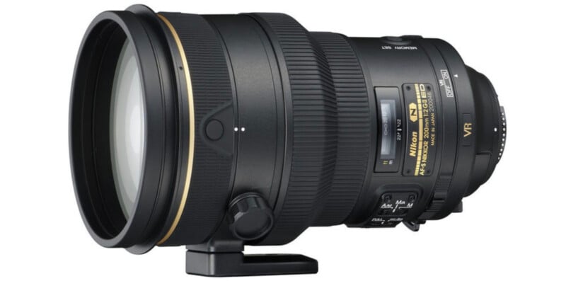 A large Nikon camera lens with a black body, gold accents, adjustment rings, and a lens mount. The lens is shown with control switches and a tripod collar attached at the base.