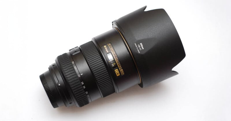 A black Nikon camera lens with a large lens hood, shown on a white background. The lens features various adjustment rings and gold lettering indicating its specifications.