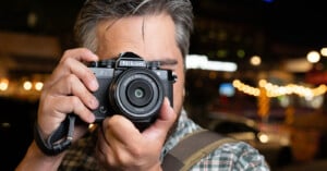 A person with dark hair and a beard holds a Nikon Zfc camera up to their face, ready to take a photo. The background is blurred with warm, glowing lights.
