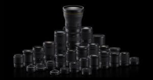 A group of various camera lenses arranged in a semi-pyramid shape on a black background, with one tall lens prominently positioned at the center back.