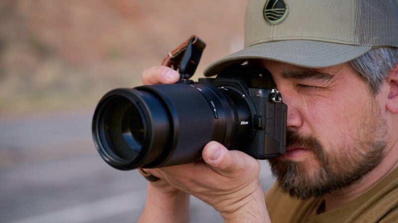 Man in a green cap taking a photo with a black DSLR camera, focusing intently. The background is blurred, suggesting an outdoor setting.