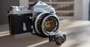 A large Nikon camera with a classic design is placed beside a miniature version on a table. The room in the background is softly lit, highlighting the intricate details of both cameras.