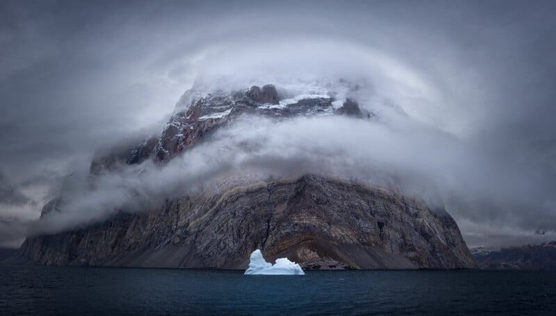 A dramatic landscape features a towering snow-capped mountain shrouded in mist, with a lone iceberg floating in the dark, calm sea beneath a cloudy, overcast sky.