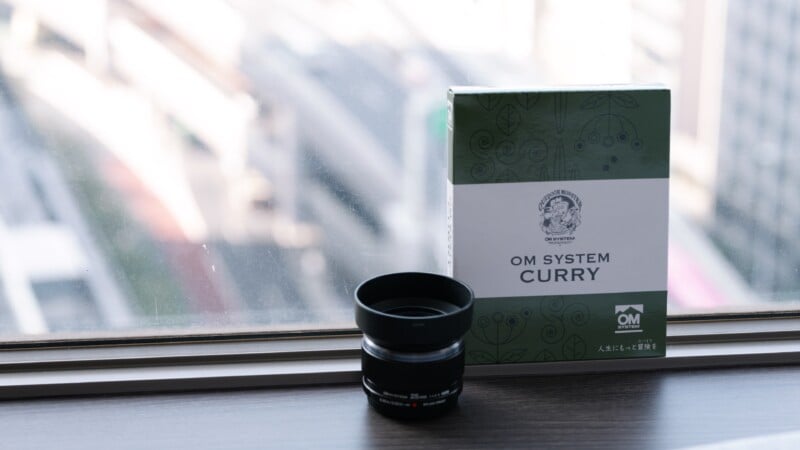 A box labeled "OM SYSTEM CURRY" with green and white design sits on a windowsill next to a camera lens. The background shows a blurred view of city buildings through the window.