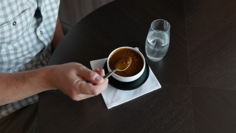 A person in a checkered shirt holds a spoonful of dark brown soup above a bowl placed on a white napkin on a dark table. A glass of water sits nearby.