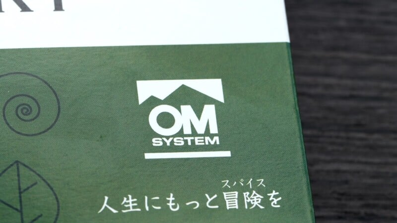Close-up of a green product packaging featuring a white logo with the text "OM SYSTEM." Japanese text is also visible, along with decorative spiral and leaf designs on a textured background.