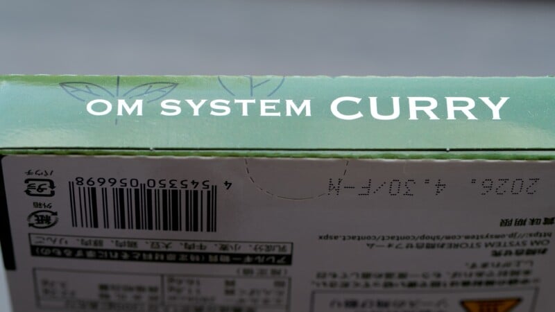 Close-up of a green box with the text "OM SYSTEM CURRY" on top. The box also features a barcode and Japanese characters. The background is blurred, and the focus is on the text and design elements on the packaging.