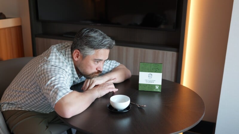 A man with gray hair and a beard is sitting at a table, resting his chin on his arms. A white cup and a small sign labeled "curry" are on the table in front of him. A TV is mounted on the wall in the background.