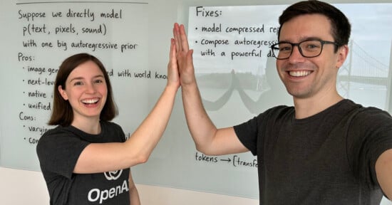 Two people smiling as they high-five in front of a whiteboard with handwritten notes. The whiteboard includes points under "Pros" and "Fixes." Both are wearing casual dark t-shirts.