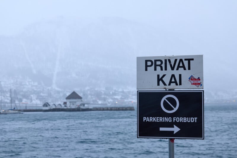 A foggy scene of a waterfront with a sign reading "Privat Kai" and "Parkering Forbudt" in Norwegian, indicating a private dock with no parking allowed. A snow-covered mountain is visible in the background.