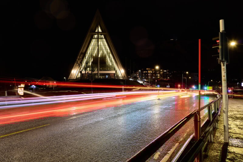 Night scene of a triangular church building with illuminated windows in the background. Streaks of red and white lights from passing vehicles on a wet road create motion blur. Lamp posts and barriers line the road.