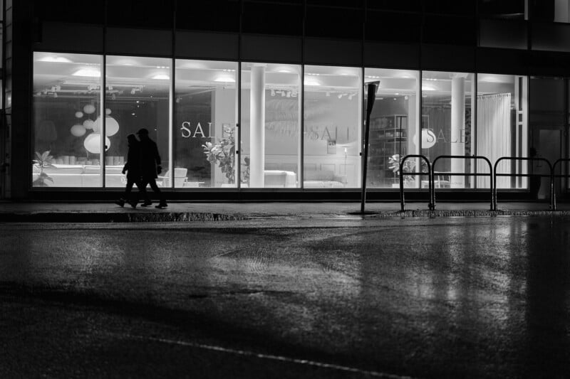 Two people walk past a brightly lit storefront displaying "SALE" signs at night. The sidewalk and street are wet, reflecting the light from the store's large windows. The atmosphere is calm and quiet.